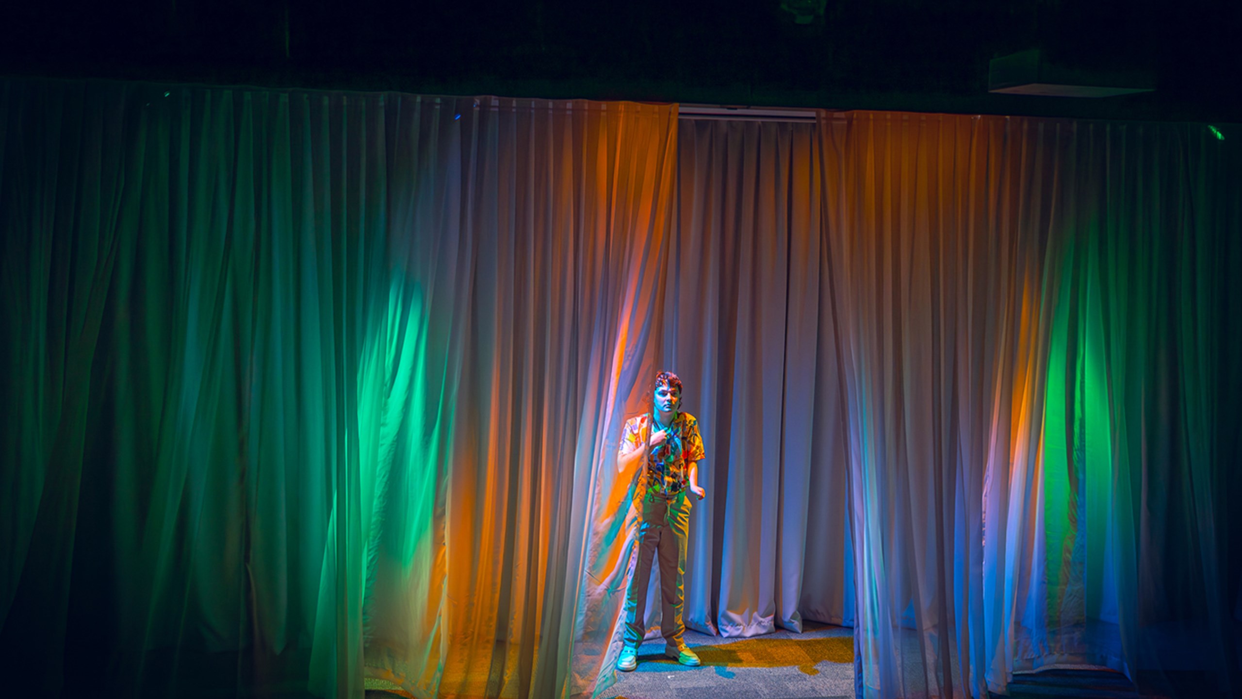 William Rees on stage surrounded by curtains which have orange, blue, and green lights projected on them